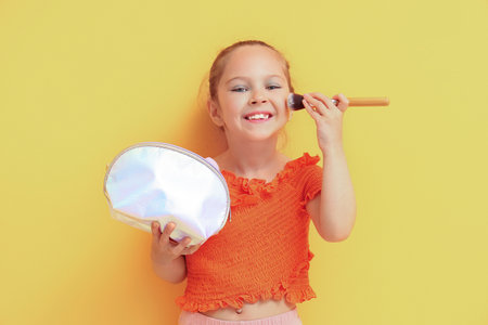 Cute little girl applying blush with cosmetic bag on yellow backgroundの写真素材