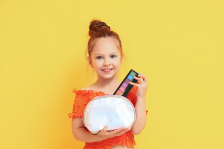 Cute little girl with cosmetic bag and palette of eyeshadows on yellow backgroundの写真素材