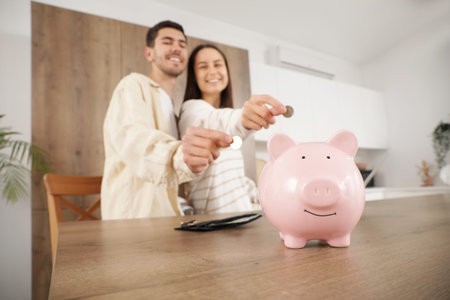 Young couple putting coins into piggy bank at table in kitchen, closeupの写真素材