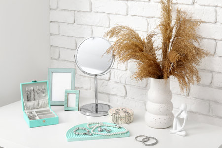 Stylish different jewelry with mirror, photo frame and vase of pampas grass on table near white brick wallの写真素材
