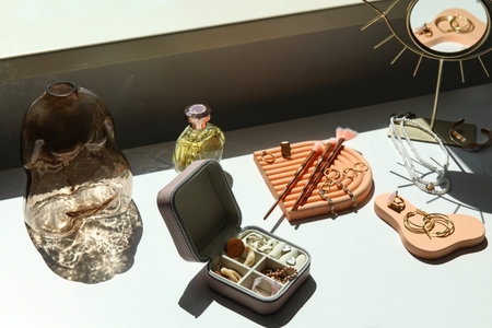 Box and stands with stylish different jewelry, mirror and perfume on table near window