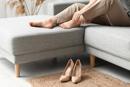 Young woman suffering from joint pain after heeled shoes on gray sofa at homeの写真素材