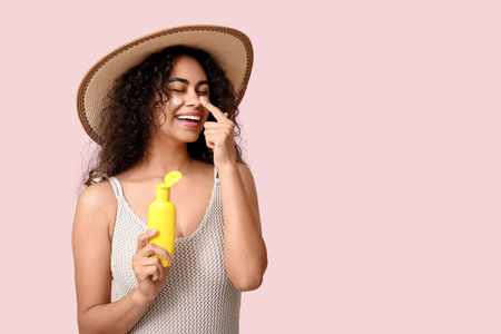 Beautiful young happy African-American woman with bottle of sunscreen cream on pink background