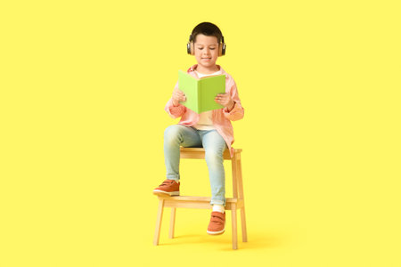 Cute little Asian boy in headphones listening to audiobook with book and sitting on stool against yellow backgroundの写真素材