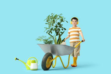Cute little Asian boy in headphones with wheelbarrow and houseplants on blue backgroundの写真素材