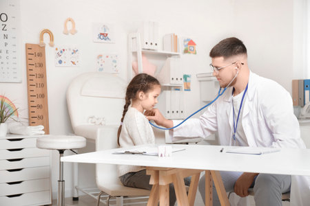 Male pediatrician with stethoscope listening to little girl at table in clinicの写真素材