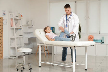 Male pediatrician examining little girl's leg on couch in clinicの写真素材