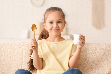 Cute little girl with spoon and yogurt sitting on sofa at homeの写真素材