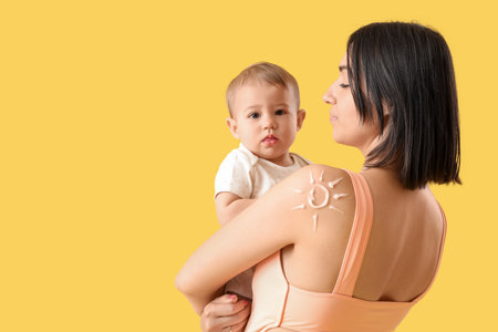 Young mother with her little baby and sun made of sunscreen cream on yellow backgroundの写真素材