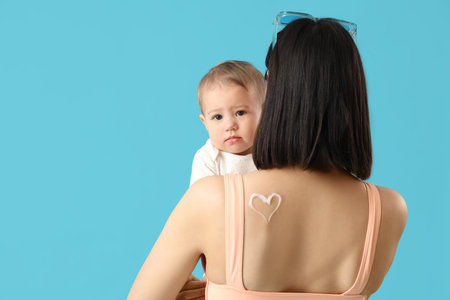 Young mother with her little baby and heart made of sunscreen cream on blue background, back viewの写真素材
