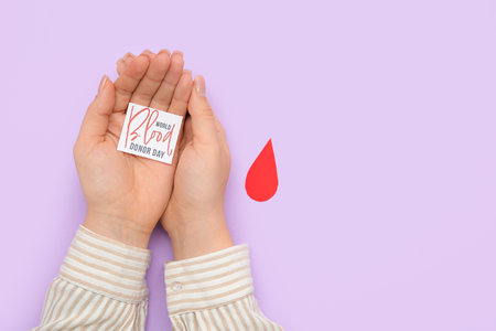Female hands holding paper piece of sheet with text WORLD BLOOD DONOR DAY and paper drop on lilac backgroundの写真素材