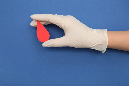 Female hand in rubber glove holding paper blood drop on blue background. Donation blood concept.の写真素材