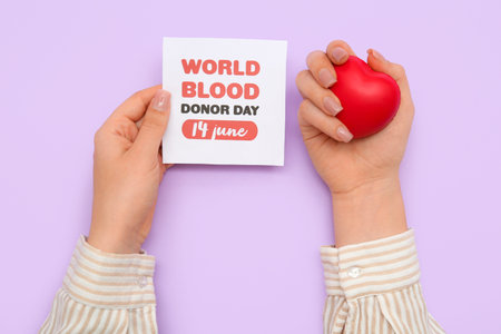 Female hands holding with grip ball and paper sheet with text WORLD BLOOD DONOR DAY 14 JUNE on lilac backgroundの写真素材