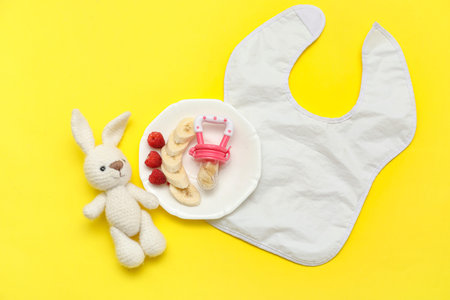 Plate with baby nibbler, fruits, toy bunny and bib on yellow backgroundの写真素材