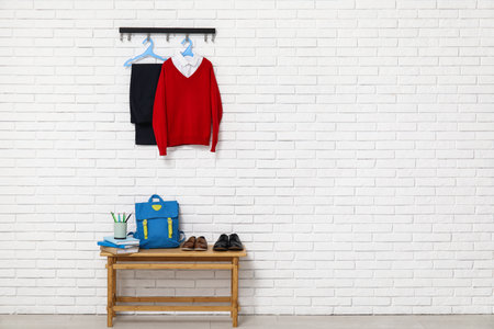 Wooden table with backpack, stationery, shoes and stylish school uniform hanging on light brick wallの写真素材