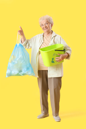 Senior woman with garbage bag and bin on yellow background. Waste sorting conceptの写真素材