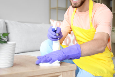 Male janitor cleaning coffee table with spray and rug in roomの写真素材
