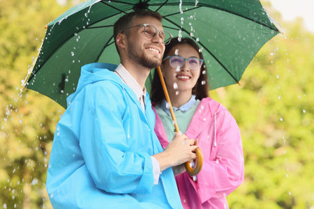 Happy young couple in raincoats holding umbrella on rainy day outdoorsの写真素材