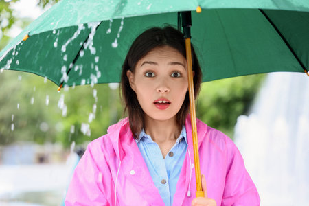 Portrait of surprised young Asian woman man in raincoat holding umbrella outdoors on rainy dayの写真素材