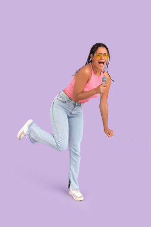 Young African-American woman in sunglasses with microphone singing on lilac background