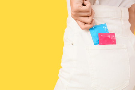 Young woman with condoms in pocket of pants on yellow background, back viewの写真素材