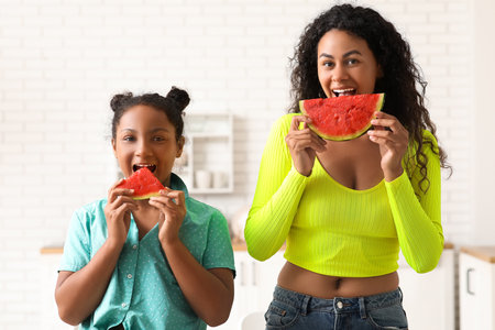 Beautiful young African-American woman and her cute daughter with pieces of fresh watermelon in kitchenの写真素材