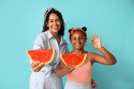 Beautiful young African-American woman and her cute daughter with slices of fresh watermelon showing ok gesture on blue backgroundの写真素材