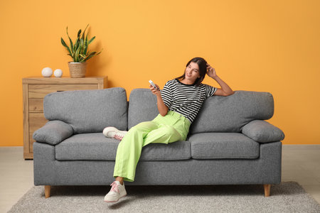Young woman with air conditioner remote control sitting on sofa near orange wall at homeの写真素材