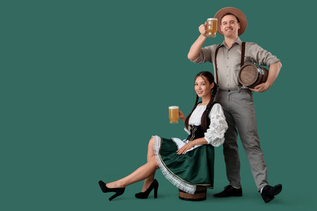 Young happy couple in traditional German clothes with barrels and glasses of beer sitting on green backgroundの写真素材