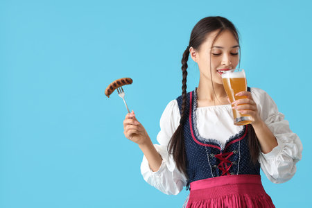 Beautiful young happy Asian Octoberfest waitress with glass of beer and sausage on blue backgroundの写真素材