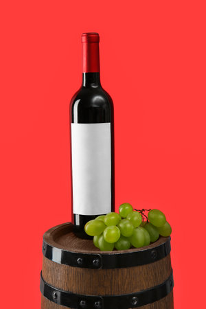 Bottle of red wine and ripe grapes on barrel against red backgroundの写真素材
