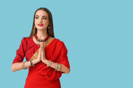 Beautiful young happy Indian woman in stylish sari praying on blue background