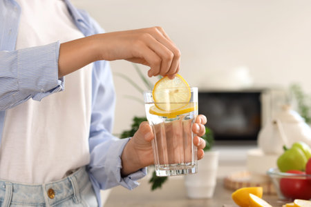 Young woman putting lemon slice into glass of water in kitchen, closeup