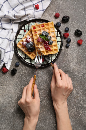 Female hands with cutlery and plate of sweet Belgian waffles with different fresh berries on gray backgroundの写真素材