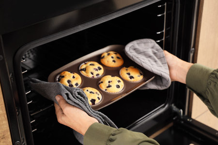 Mature woman taking baking dish with muffins from oven in kitchen, closeup
