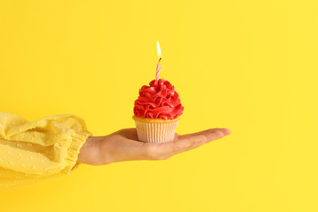 Woman holding sweet Birthday cupcake with burning candle on yellow backgroundの写真素材