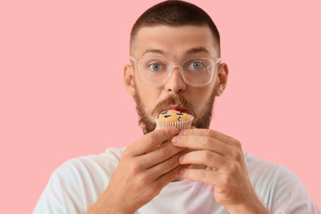 Shocked young man eating tasty muffin on pink background, closeup