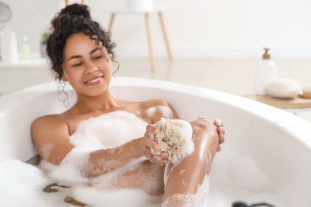 Beautiful young happy African-American woman taking bath with sponge and soap foam at homeの写真素材