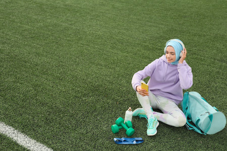 Sporty Muslim woman in headphones with sport equipment and mobile phone sitting on fake grass at stadiumの写真素材