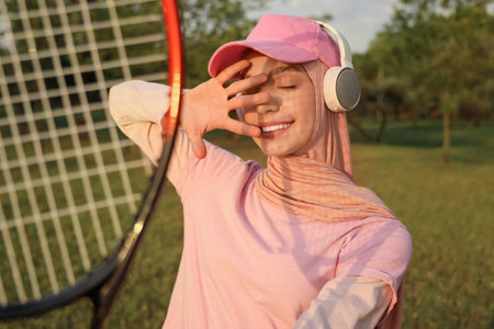 Sporty Muslim woman in headphones with tennis racket in parkの写真素材