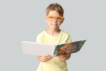 Cute little boy in eyeglasses reading book on gray backgroundの写真素材