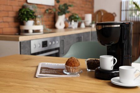 Modern coffee machine with cups, beans and muffin on table in kitchen