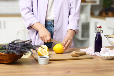 Young woman cutting lemon for preparing lavender lemonade at table in kitchen