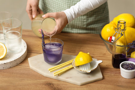 Young woman pouring lemon juice from jug into glass for preparing lavender lemonade at table in...