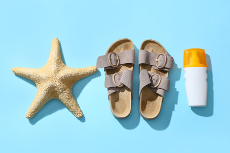 Stylish sandals with starfish and bottle of sunscreen cream on blue background