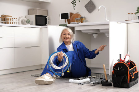 Mature female plumber with pipes pointing at sink in kitchen