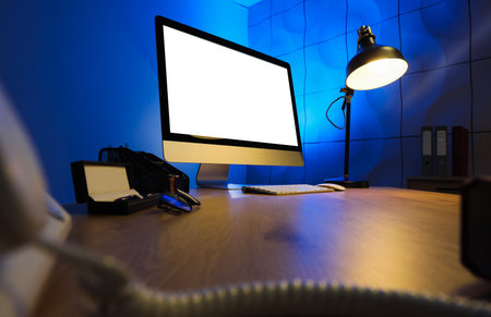 Blank computer monitor with glowing lamp on desk in dark office