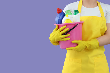 Female janitor with bucket of cleaning supplies on lilac background, closeupの写真素材