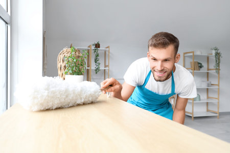 Male janitor with dust brush cleaning furniture in light room