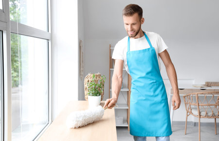 Male janitor with dust brush cleaning furniture in light room